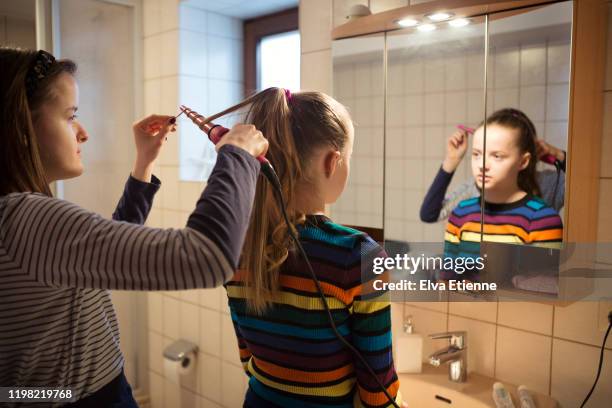 teenage girl using hair curling tongs on younger sister's hair in a domestic bathroom - domestic room imagens e fotografias de stock