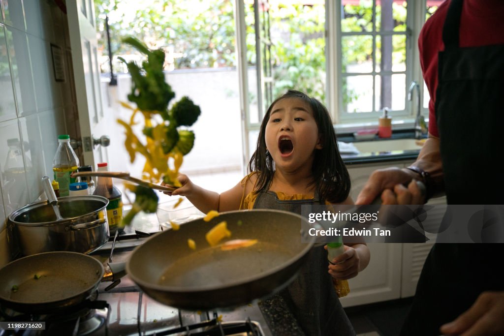 An Asian Chinese female girl looking at her father cooking in the kitchen preparing food for her and learning