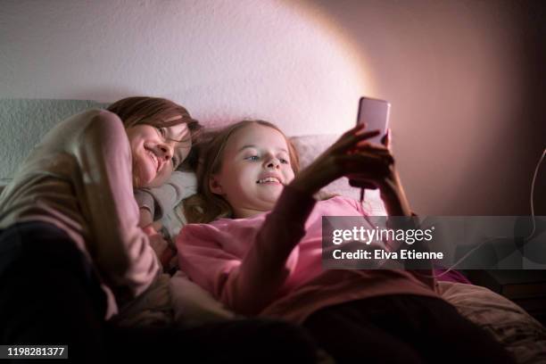 two adolescent sisters using a mobile phone plugged in to a power supply, lying on a bed at night - electrical component stock pictures, royalty-free photos & images