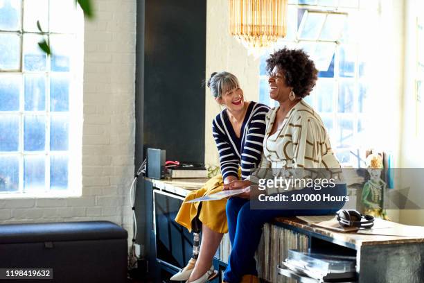 mature women laughing together in stylish loft apartment - vrouw 50 jaar stockfoto's en -beelden