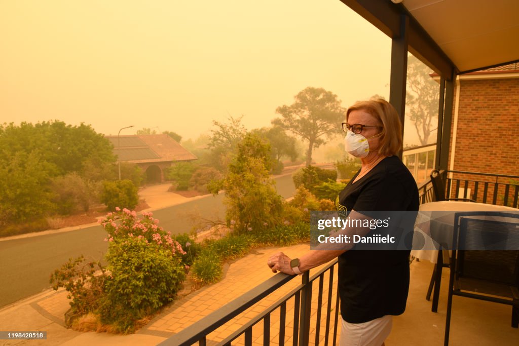A woman, on a balcony, wears a face mask to protect herself against very dangerous level of air pollution.  A thick yellow fog coming from the nearby forest fires covers Canberra city.