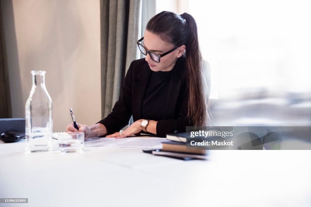 Businesswoman writing on documents while sitting by desk at law firm