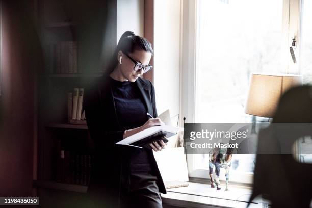female professional writing on documents while standing by window at law firm - finanzwirtschaft und industrie stock-fotos und bilder