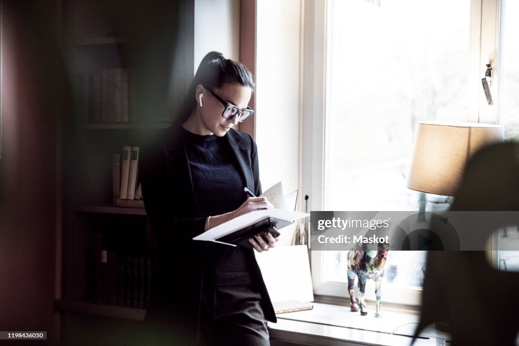 Female professional writing on documents while standing by window at law firm