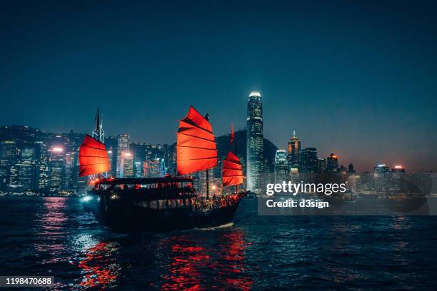 traditional chinese junk boat sailing across victoria harbour against illuminated hong kong city skyline at night - hong kong junk boat stock-fotos und bilder