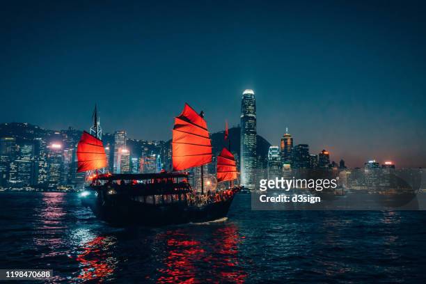 traditional chinese junk boat sailing across victoria harbour against illuminated hong kong city skyline at night - hong kong junk boat stock pictures, royalty-free photos & images