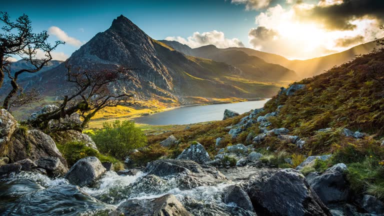 Crane shot of landscape at Snowdonia National Park in Wales. Mountain stream with fresh water in the foreground.
Tryfan mountain and Llyn Ogwen in the background. Warming evening sunlight producing sunbeams in the distance. Northern Wals, UK