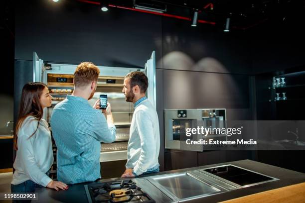 store salesman taking a snapshot of a modern designer fridge on a smart phone with an interested husband and wife standing together next to it in a kitchen equipment store - household equipment stock pictures, royalty-free photos & images