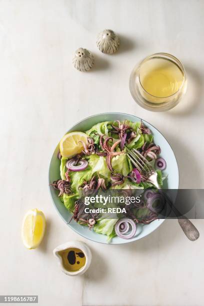 Green salad with grilled squids calamari tentacles, lemon, purple onion in ceramic bowl, serving with olive oil sauce, glass of white wine over white...