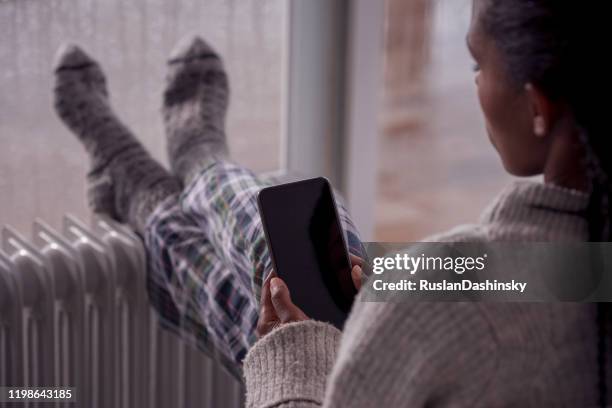 eine frau heizt sich die beine vor einer elektrischen heizung auf, während sie ein smartphone benutzt. - müdigkeit winter stock-fotos und bilder