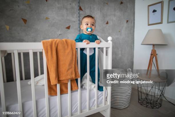 verspielte baby junge mit schnuller stehen in krippe nach dem aufwachen - babybett stock-fotos und bilder