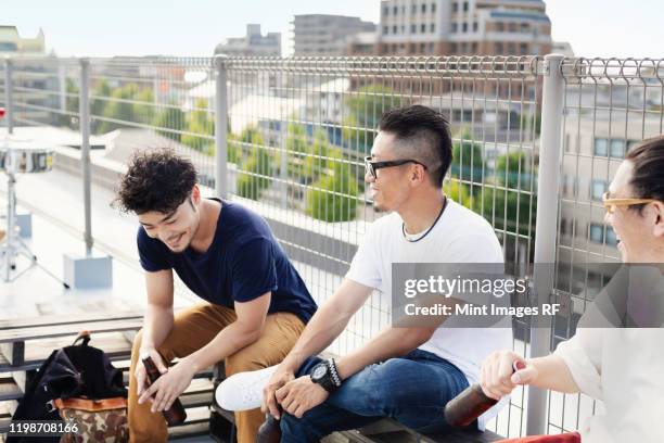 three young japanese men sitting on a rooftop in an urban setting, drinking beer. - man sipping beer smiling stockfoto's en -beelden