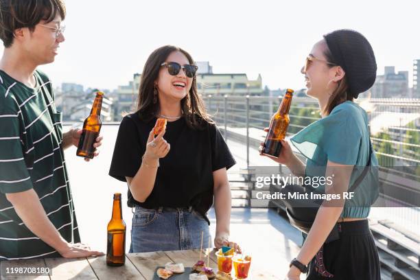 young japanese man and two women standing on a rooftop in an urban setting, drinking beer. - 日本人　女性　友達 ストックフォトと画像