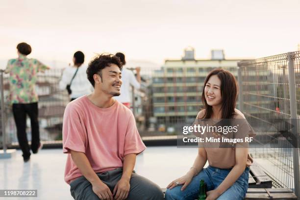 smiling young japanese man and woman sitting on a rooftop in an urban setting. - trend ストックフォトと画像