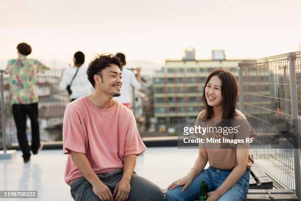 smiling young japanese man and woman sitting on a rooftop in an urban setting. - youth culture guy stock pictures, royalty-free photos & images