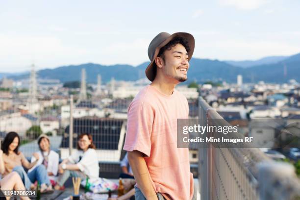 smiling young japanese man standing on a rooftop in an urban setting. - hats ストックフォトと画像