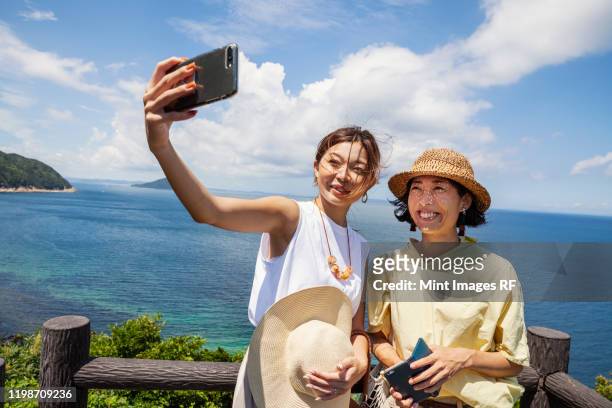 two japanese women wearing hats standing on a cliff, taking selfie with mobile phone, ocean in the background. - autorretratarse fotografías e imágenes de stock