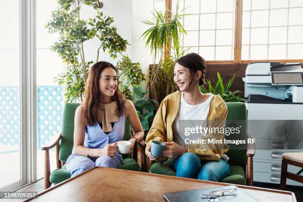 two female japanese professionals sitting in a co-working space, smiling at each other. - creative rf stock-fotos und bilder