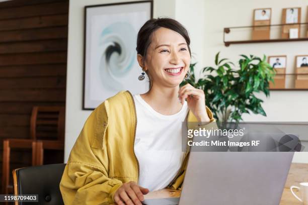 female japanese professional sitting at a table in a co-working space, using laptop computer. - japanese woman stock pictures, royalty-free photos & images
