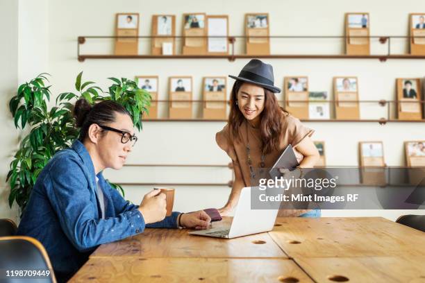 male and female japanese professional at a table in a co-working space, using laptop computer. - rf business stock pictures, royalty-free photos & images