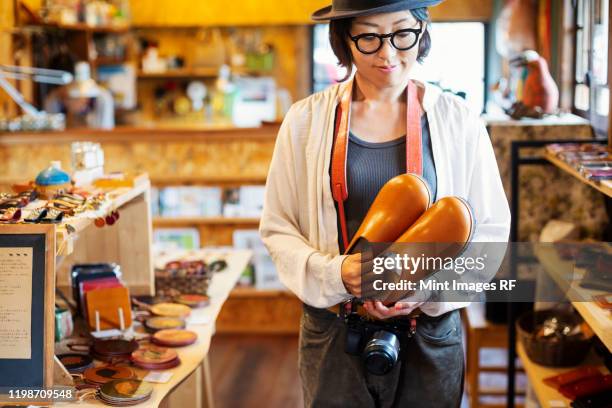 japanese woman wearing hat and glasses standing in a leather shop, holding pair of leather shoes. - leather shoe stock pictures, royalty-free photos & images