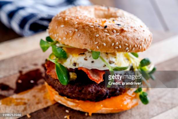 bagel do pequeno almoço com ovo, verdes, abacate, bacon, salsicha e queijo em uma placa de corte rústica - muffin inglês - fotografias e filmes do acervo