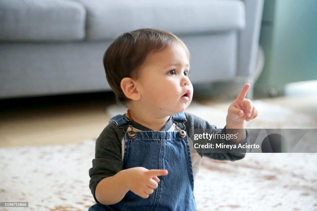 A LatinX toddler points his index finger, while sitting in his living room wearing jean overalls and a long sleeved t-shirt.