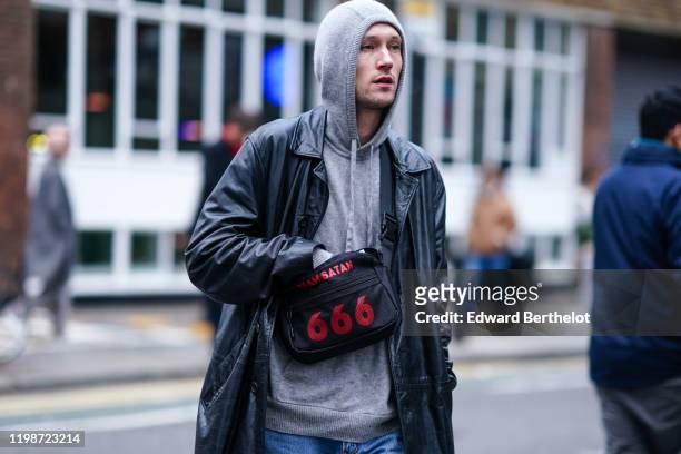 Guest wears a gray hoodie sweater, a black leather long coat, a fanny pack bag with printed " Team Satan 666", during London Fashion Week Men's...