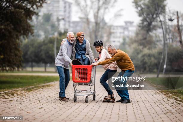 playful senior couples having fun with shopping cart and skateboard in the park. - man pushing cart fun play stock pictures, royalty-free photos & images