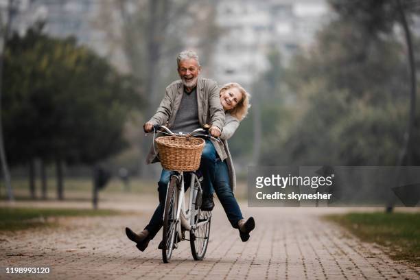 coppia senior giocosa che si diverte in bicicletta nella giornata autunnale. - coppia anziana foto e immagini stock