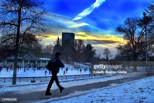 Cold winter sun goes down behind the Hancock Building as a bundled-up pedestrian walks by the Frog Pond Pavilion on Boston Common, which is filled...