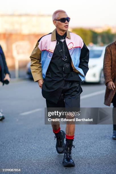 Evan Mock, skateboarder, wears sunglasses, a Prada jacket, a black Prada shirt, a mini bag, black shorts, red and black socks, leather shoes, outside...