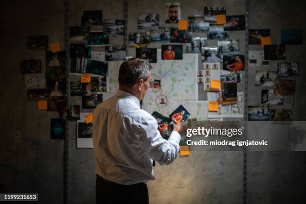 mature police detective looking at pictures while standing in front of the wall - police station imagens e fotografias de stock