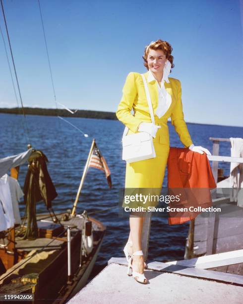 Esther Williams, US actress and former swimmer, wearing a yellow dress and carrying a white handbag as she poses by the water's edge, alongside a...