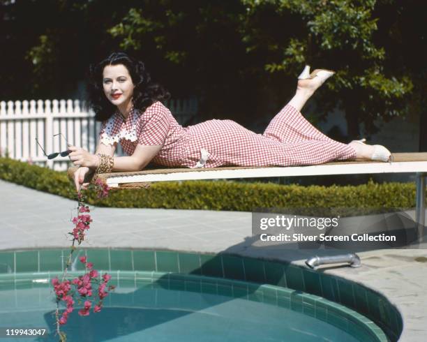 Hedy Lamarr , Austrian actress, wearing a red gingham dress and holding a branch with pink flowers while leaning, front down, on a diving board over...