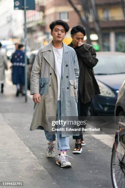 Guest wears a gray and pale blue trench coat with large pockets, blue cuffed jeans, a white t-shirt, a necklace, socks, sneakers shoes, outside...