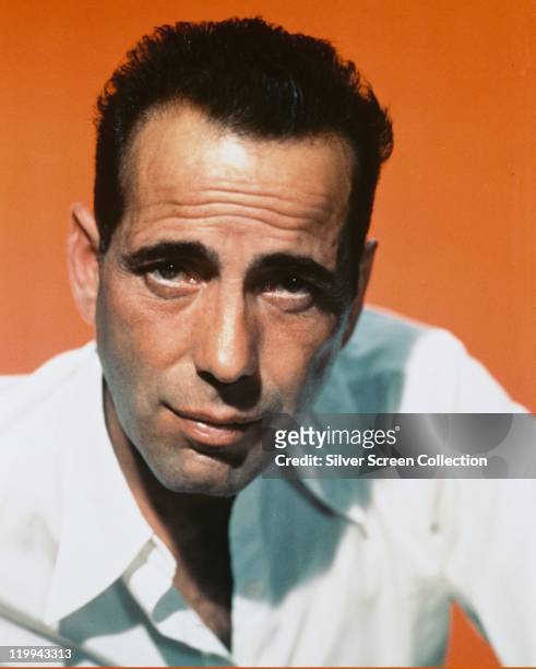 Headshot of Humphrey Bogart , US actor, wearing a white open-necked shirt in a studio portrait, against an orange background, circa 1940.