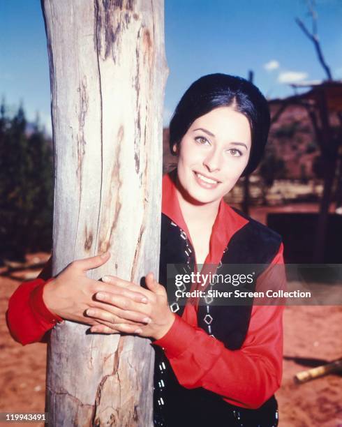 Ina Balin , US actress, wearing a red blouse, beneath a black waistcoat, hugging a tree trunk, circa 1970.