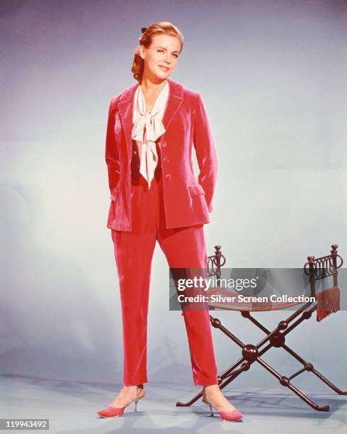 Full-length shot of Honor Blackman, British actress, weaing a red trouser suit, white blouse and red shoes, posing beside a chair in a studio...