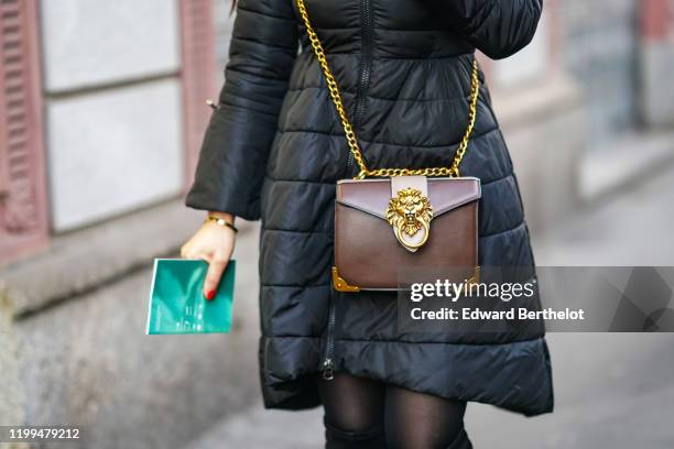 Guest wears a black long winter puffer coat, a brown bag with a metallic golden part depicting a lion's head, the bag is attached to a golden chain,...