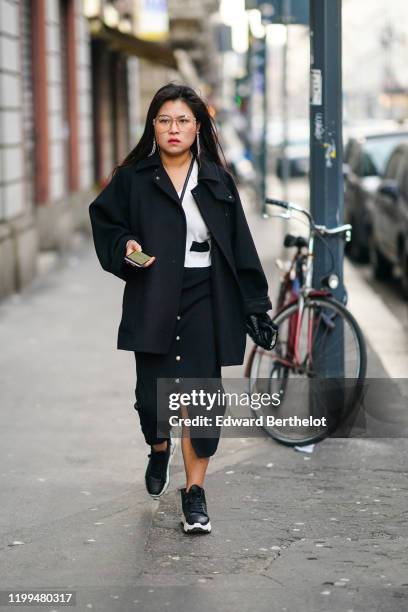 Guest wears glasses, earrings, a black jacket, a white top, a skirt with buttons, black sneakers shoes, outside Reshake, during Milan Fashion Week...