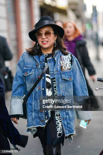 Guest wears a blue denim ripped jacket with printed "The Bad Guy", sunglasses, earrings, a hat outside Reshake, during Milan Fashion Week Menswear...