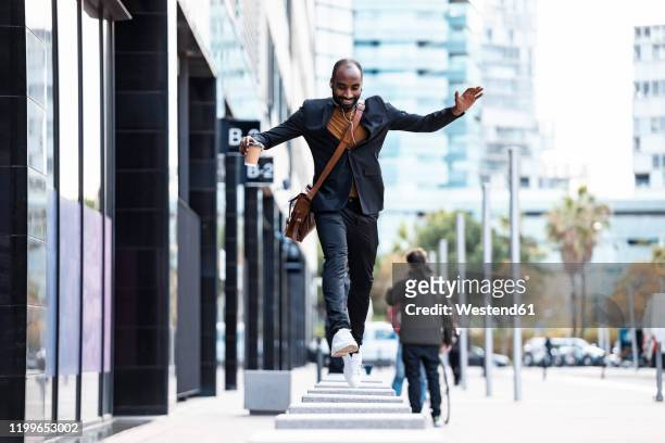 young businessman with earphones and coffee to go jumping from bollard to bollard - business people on the move stock pictures, royalty-free photos & images