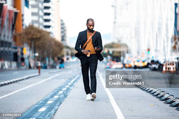 young businessman with coffee to go looking at cell phone while walking on the street - black coat stock pictures, royalty-free photos & images