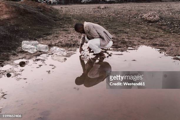 young man filling plastic bottle with water at a water hole - schmutzig stock-fotos und bilder