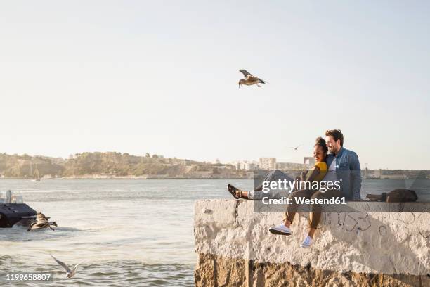 young couple sitting on pier at the waterfront enjoying the view, lisbon, portugal - tourism in lisbon stock pictures, royalty-free photos & images