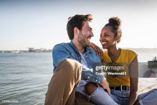 happy young couple sitting on pier at the waterfront, lisbon, portugal - steg zwei menschen stock-fotos und bilder