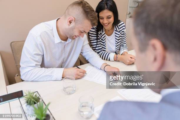 junges glückliches paar unterschreibt ein dokument, während er sich mit seinem immobilienmakler im büro trifft - alliance mariage stock-fotos und bilder