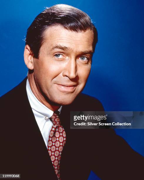 James Stewart , US actor, weaing a black jacket, white shirt and red patterned tie in a studio portrait, against a blue background, circa 1955.