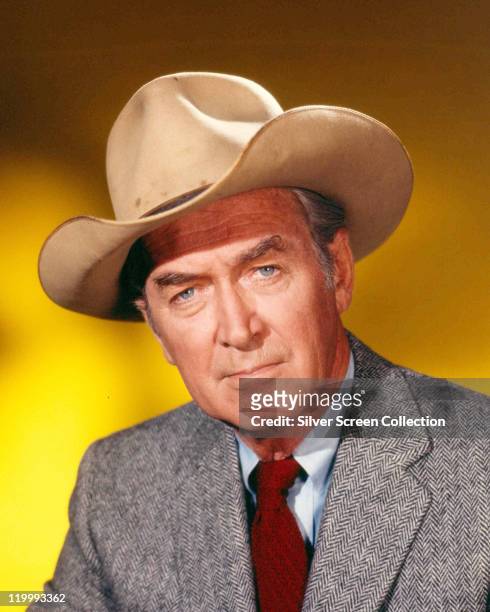 James Stewart , US actor, wearing a cowboy hat, a grey herringbone jacket, white shirt and red tie in a studio portrait, against a yellow background,...